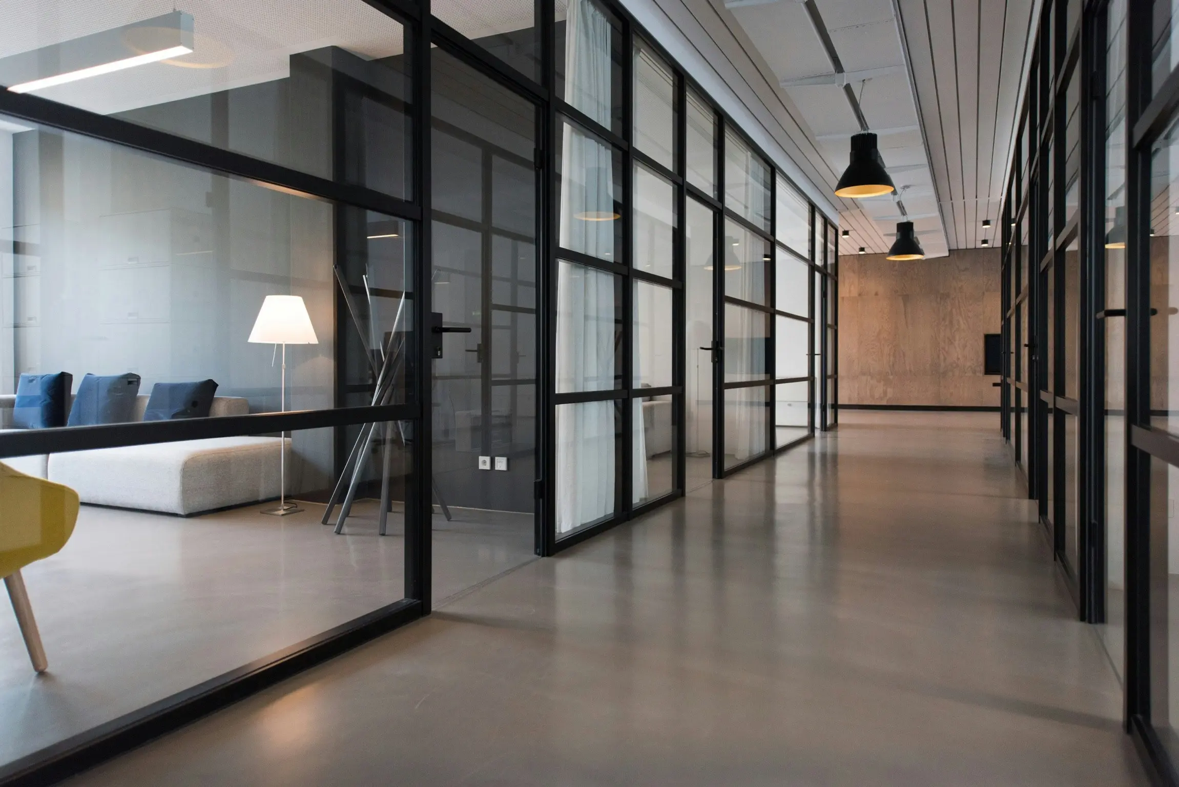 Pasillo de oficina moderno con salas de reuniones con paredes de vidrio, piso pulido, un sofá blanco y una lámpara a la izquierda y luces de techo negras.