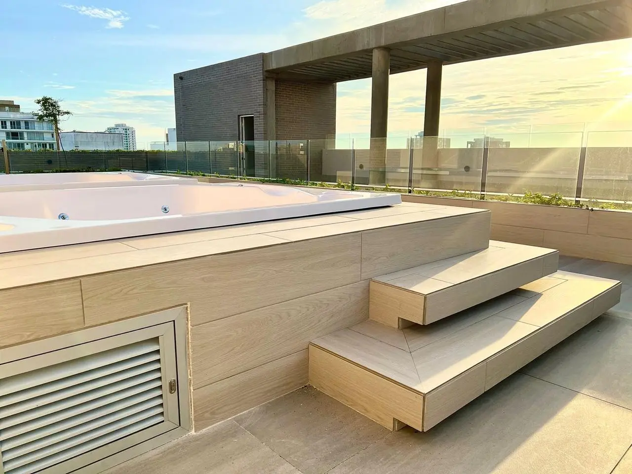 Un tranquilo espacio en la azotea con una bañera de hidromasaje, elegantes escalones de madera y una barandilla de cristal con el cielo despejado como telón de fondo.