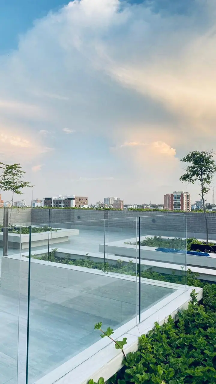 Un espacio moderno en la azotea con barandillas de cristal, vegetación y una vista panorámica de los edificios bajo un cielo azul con nubes suaves.