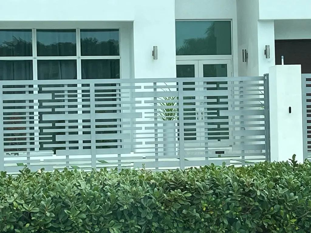 Fachada moderna de una casa con una elegante valla metálica y vegetación en la parte delantera, con grandes ventanas y elementos de diseño minimalista.