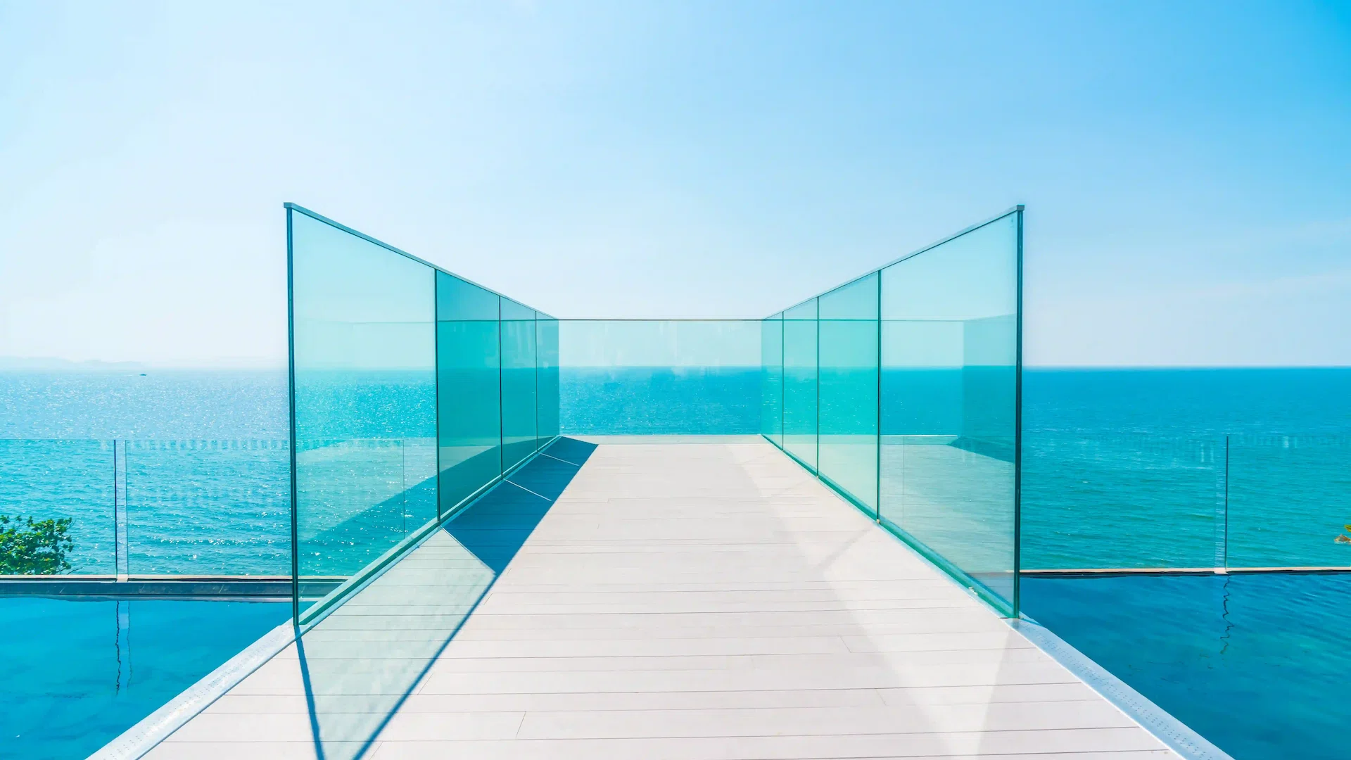 Una elegante barandilla de vidrio enmarca una terraza de madera con vista a un sereno océano azul bajo un cielo despejado, creando una sensación de tranquilidad moderna.